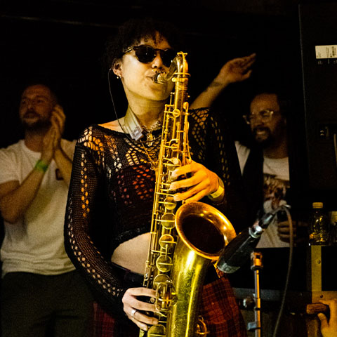 a woman playing the saxophone at a gig with two dancing men behind her