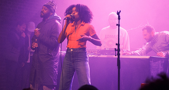 a woman performing on stage with others controlling the sound in the background