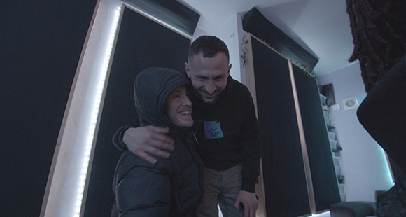 two men smiling and embracing with windows covered with black blinds